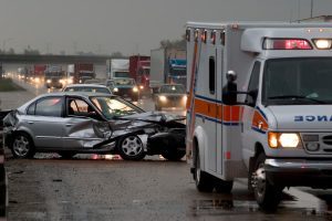 Damaged silver sedan after car crash with ambulance in foreground and traffic in background